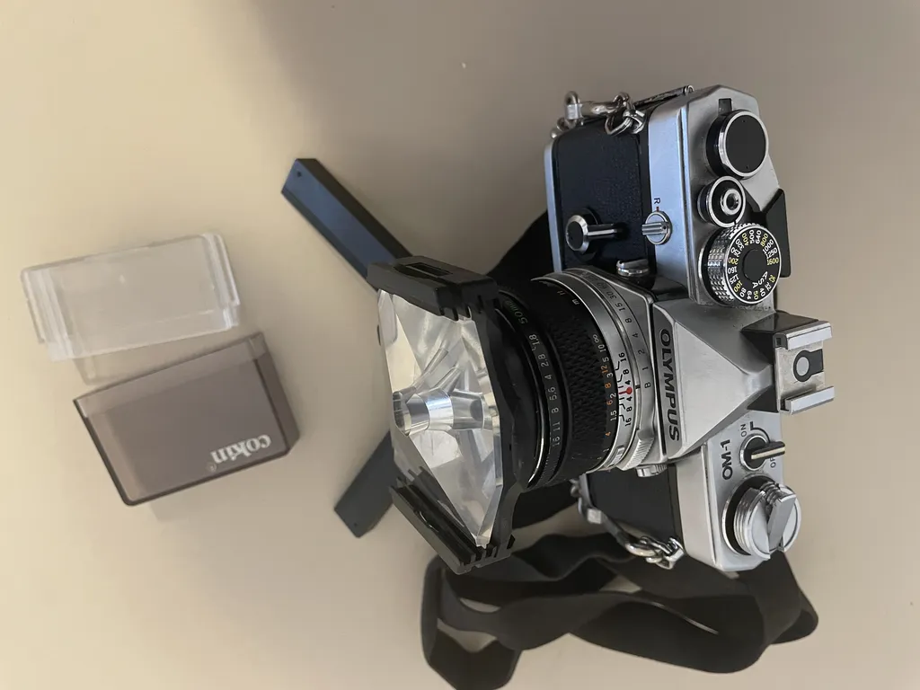 a black and silver metal camera on a tiny tripod. On the front of the lens is a black piece of plastic with ridged slots on either side. a thick, angular piece of plastic with a hole in the center is slotted in