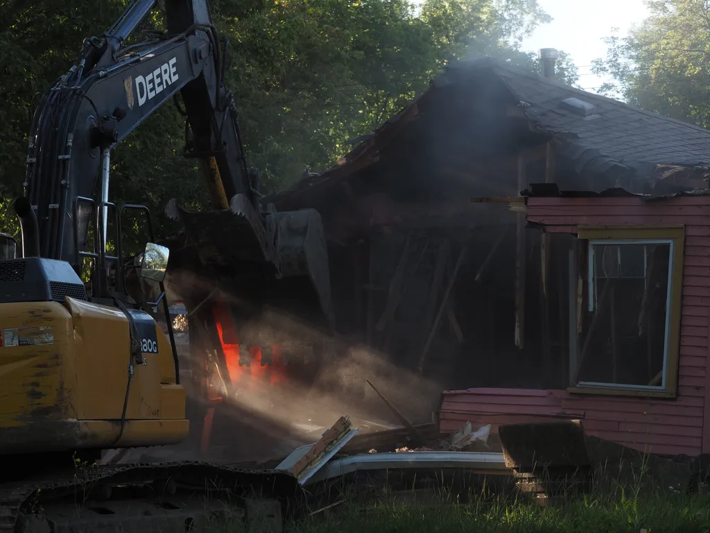 an excavator tearing down a one-story house. rays of light are catching in the dust and an orange tarp is lit up like a fire