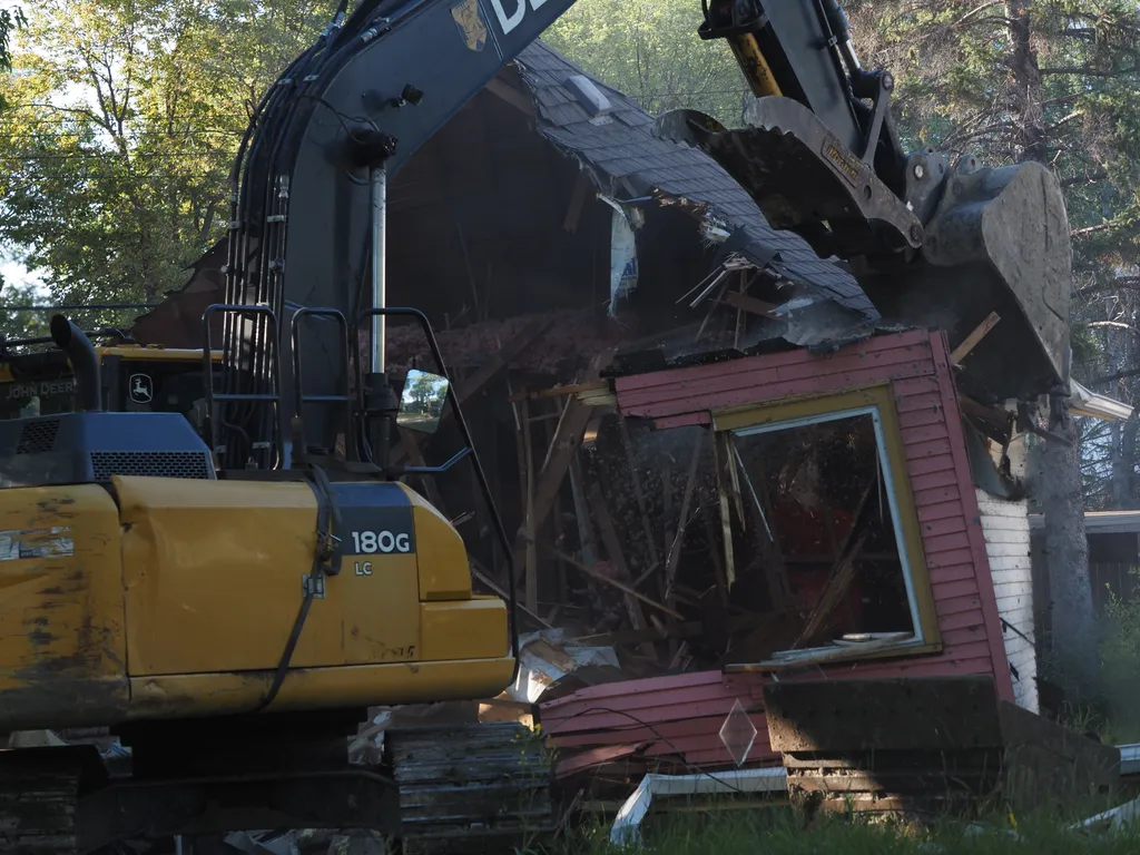 an excavator tearing down a small house. glass and debris are in the air