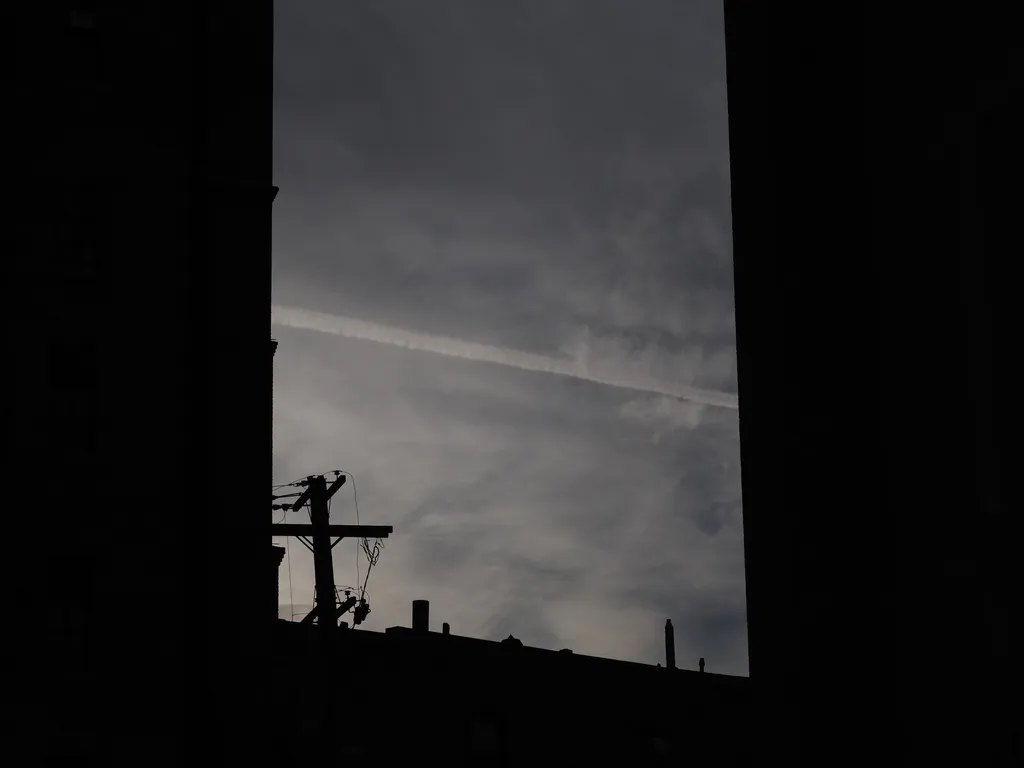 a contrail in a sky full of wispy clouds, as viewed between the silhouettes of tall brick buildings