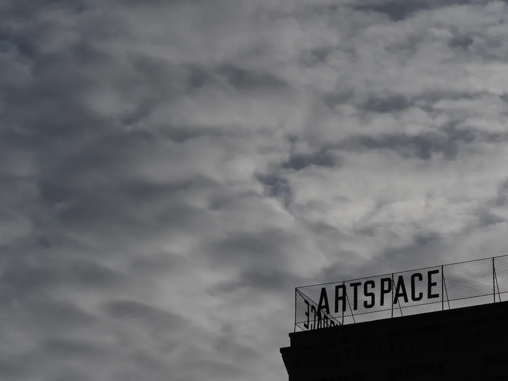 the top of a brick building silhouetted against a cloudy sky. On top of the building is a sign with letters written against a mesh background that reads 'artspace' in all-caps