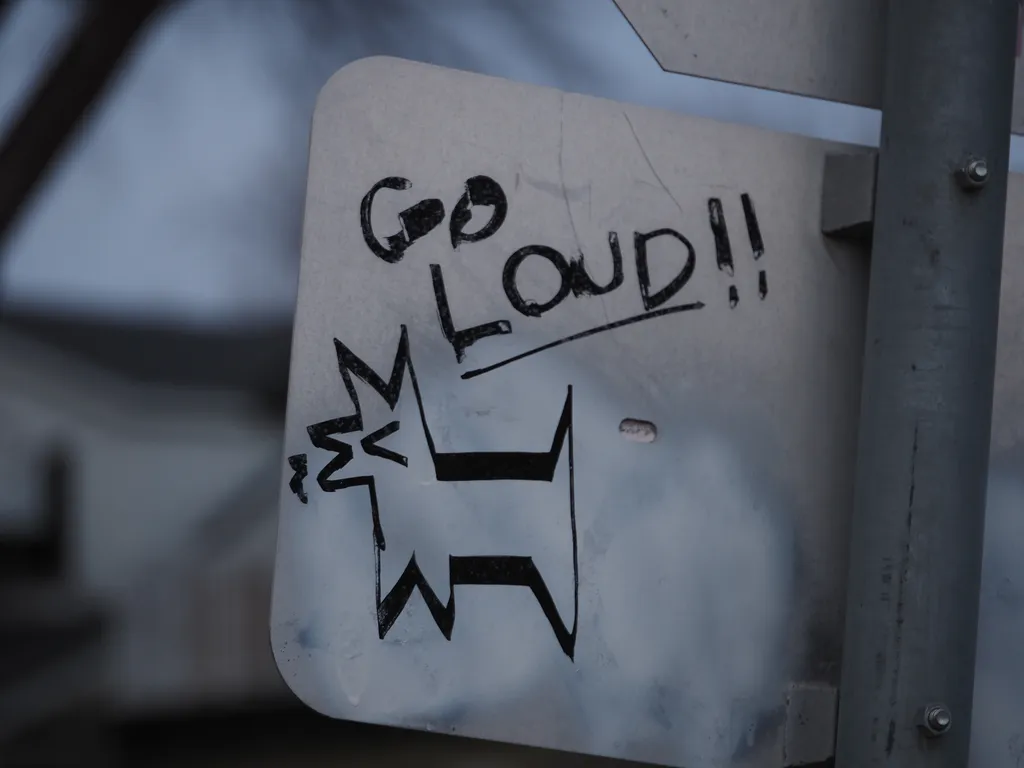 an angular dog drawn onto the back of a street sign with the accompanying message: Goo Loud!!