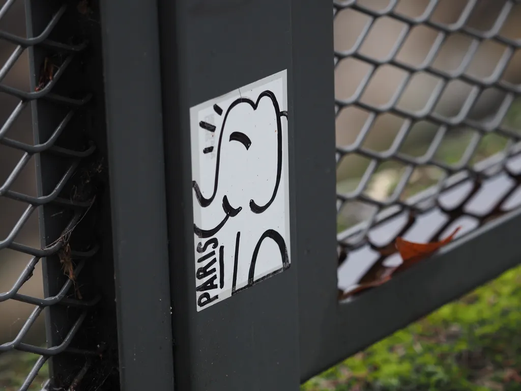 a sticker attached to a metal fence featuring the word 'Paris' and a smiling elephant drawn with minimal strokes