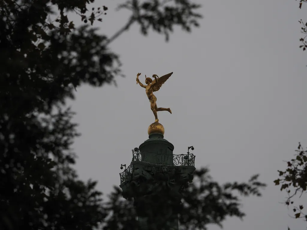 A golden statue of an angel holding a torch (the spirit of freedom) on a tall column as viewed through branches