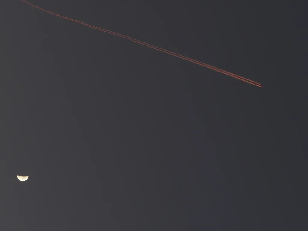 a contrail lit up by a setting sun by a quarter moon