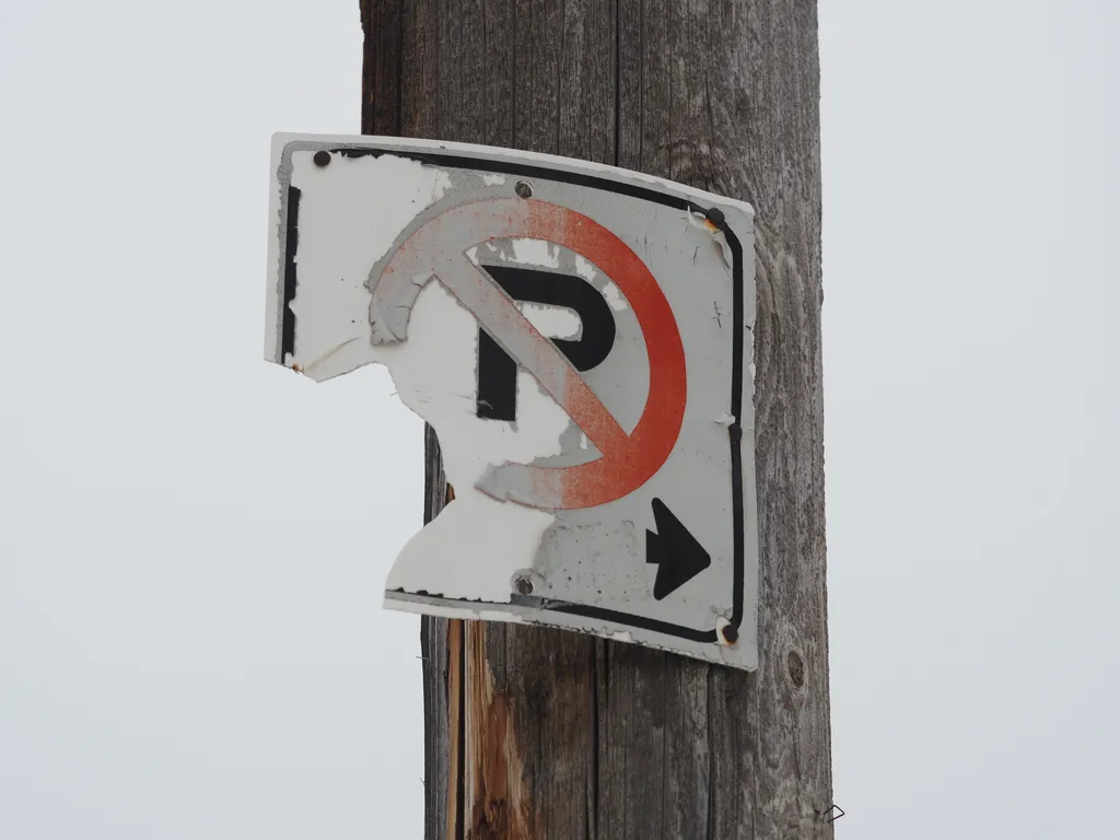 a no parking sign that has become partially unmoored from the telephone pole it's attached to and with half of its paint peeled off