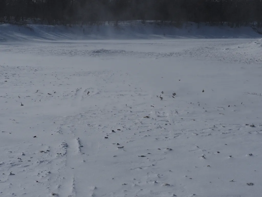 a frozen river with a path faintly visible being covered in blowing snow