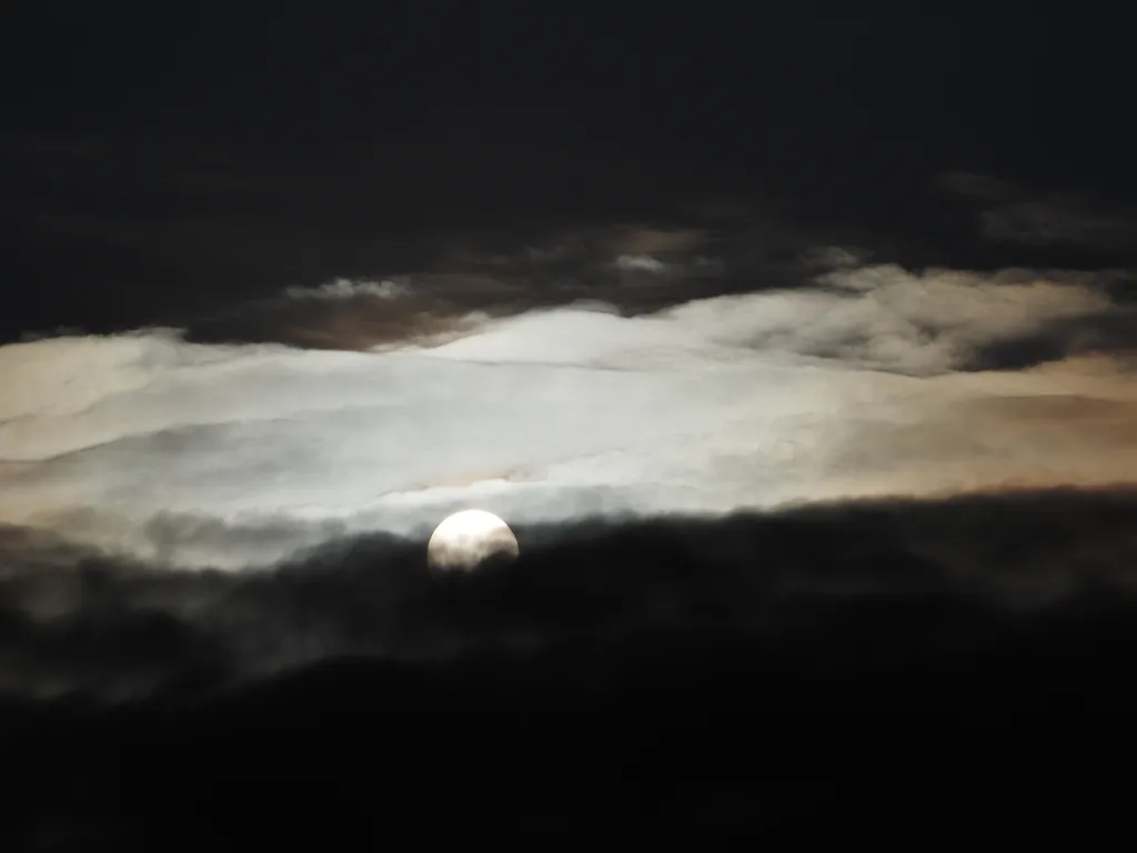 a bright sun setting behind a black cloud, with a white cloud and a black sky behind it