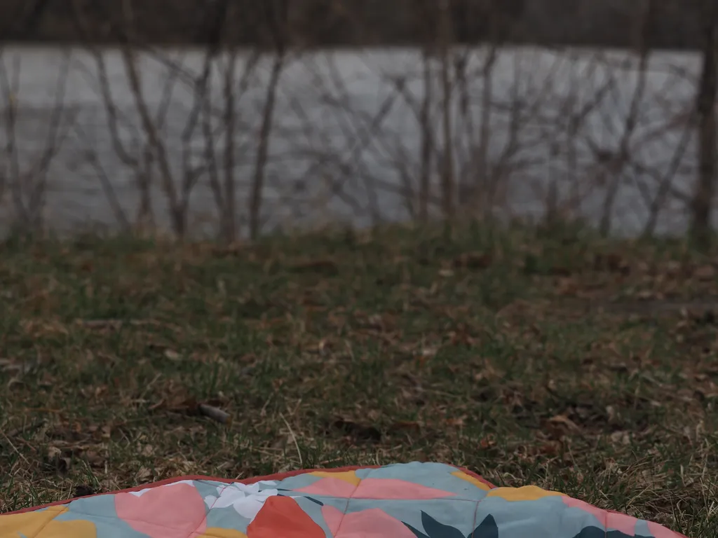 the edge of a floral picnic blanked visible on a grassy riverbank looking over an out-of-focus river