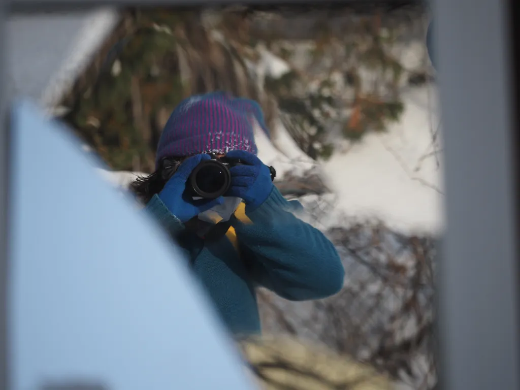 Our intrepid photographer, wearing a thick sweater, gloves, and a toque, and with a camera obscuring their face, reflected in a broken mirror